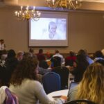 a crowd of attendees at the 2022 CHeP community engagement day views keynote speaker on the screen at the head of the room