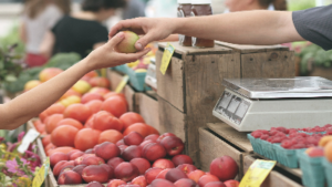 photo of hands passing fruit to one another with a farmers market in the background
